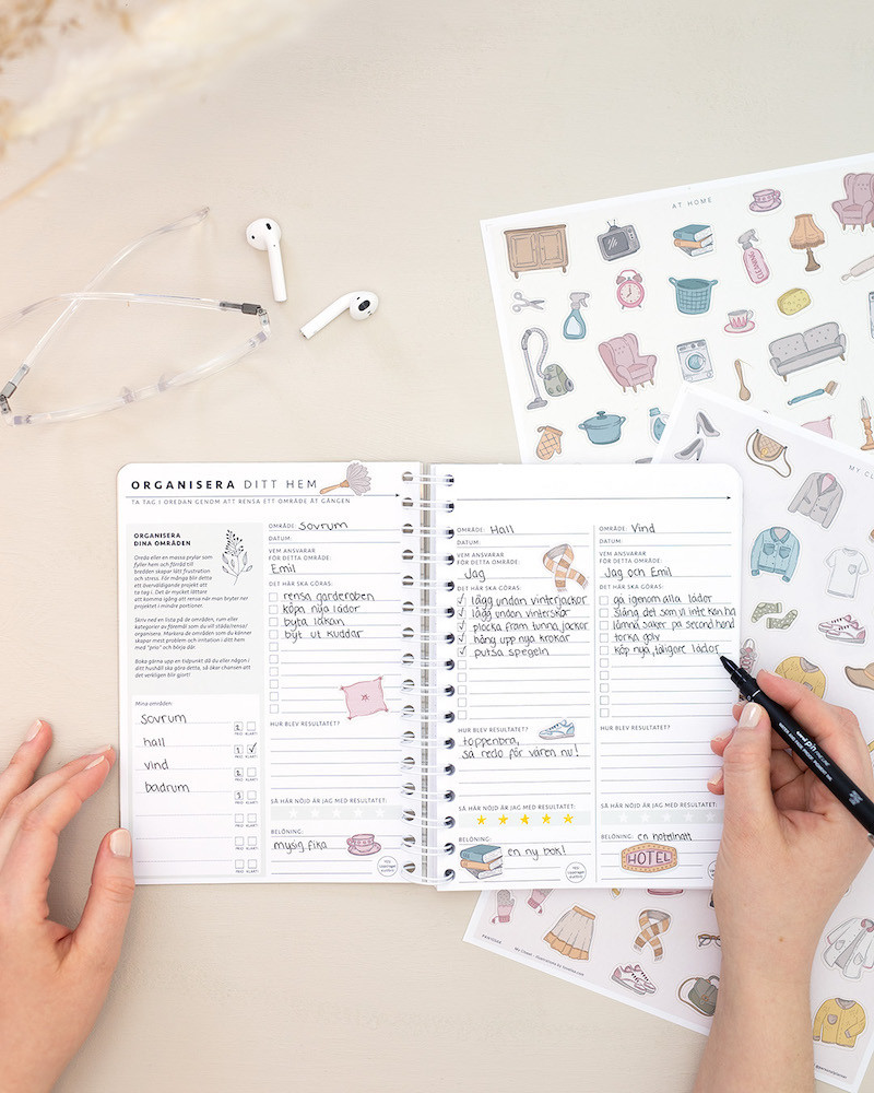 Temasidan Organisera ditt hem uppslagen i en almanacka. En hand hller i en Fineliner Uni Pin-penna. Bredvid ligger klistermrken frn arken My Closet och At home