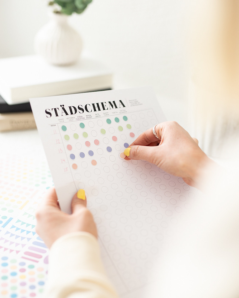 Städschemat utskriven som freebie. En hand fyller i Städschemat genom att fästa klistermärken för att bocka av uppgifter