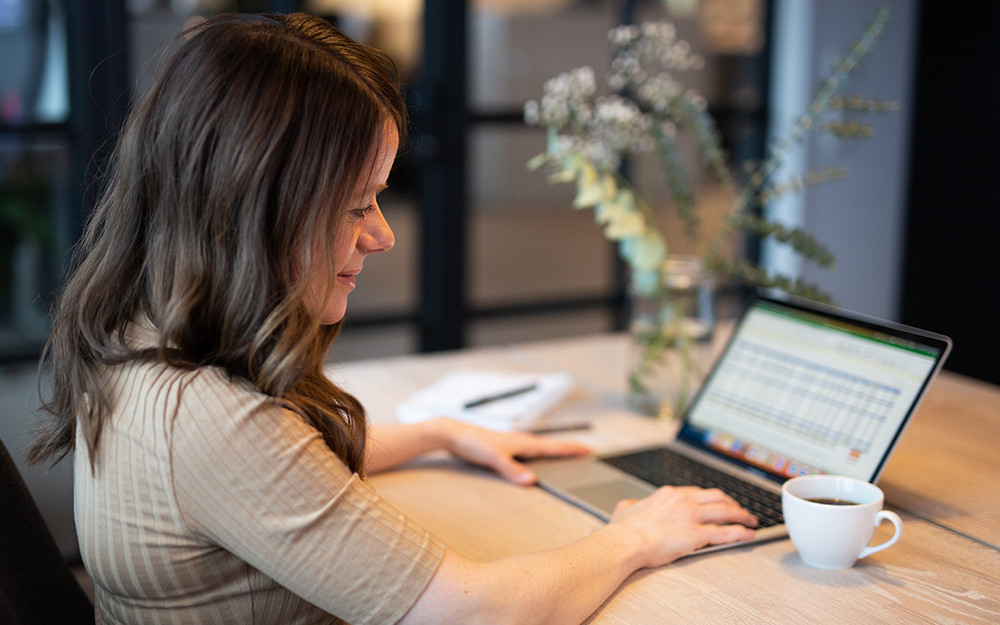 En kvinna som sitter framför en uppslagen laptop med en kaffekopp bredvid